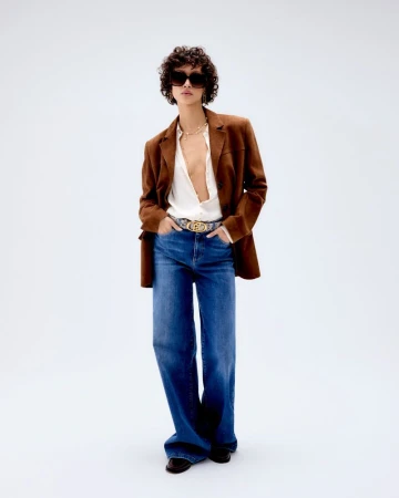 A stylish adult in sunglasses, a brown jacket, jeans, and a white top poses in front of a white background, presenting a fashion-forward look.