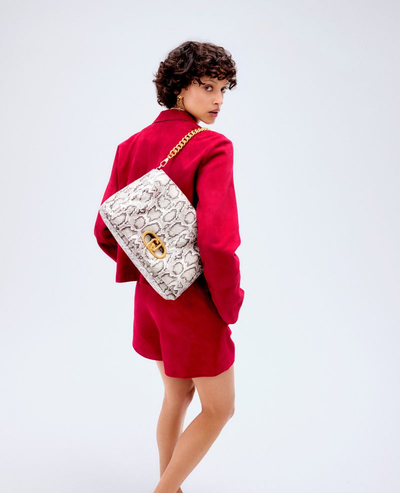 Brunette woman in a red short set poses in a studio with a patterned handbag and a chain strap slung over her shoulder.