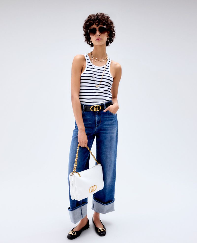 Full-length shot of a woman wearing a striped tank top, jeans, sunglasses, jewelry, and flats, holding a white purse against a white backdrop.