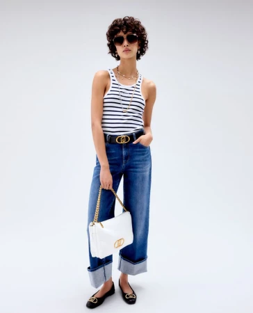 Full-length shot of a woman wearing a striped tank top, jeans, sunglasses, jewelry, and flats, holding a white purse against a white backdrop.