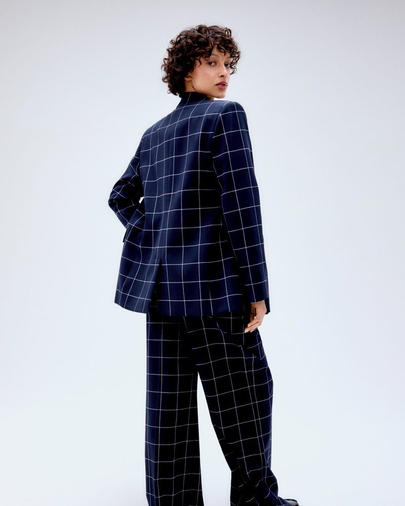 Adult woman in a blue plaid suit poses in a studio, looking over her shoulder, with curly hair and a neutral facial expression.