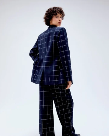 Adult woman in a blue plaid suit poses in a studio, looking over her shoulder, with curly hair and a neutral facial expression.