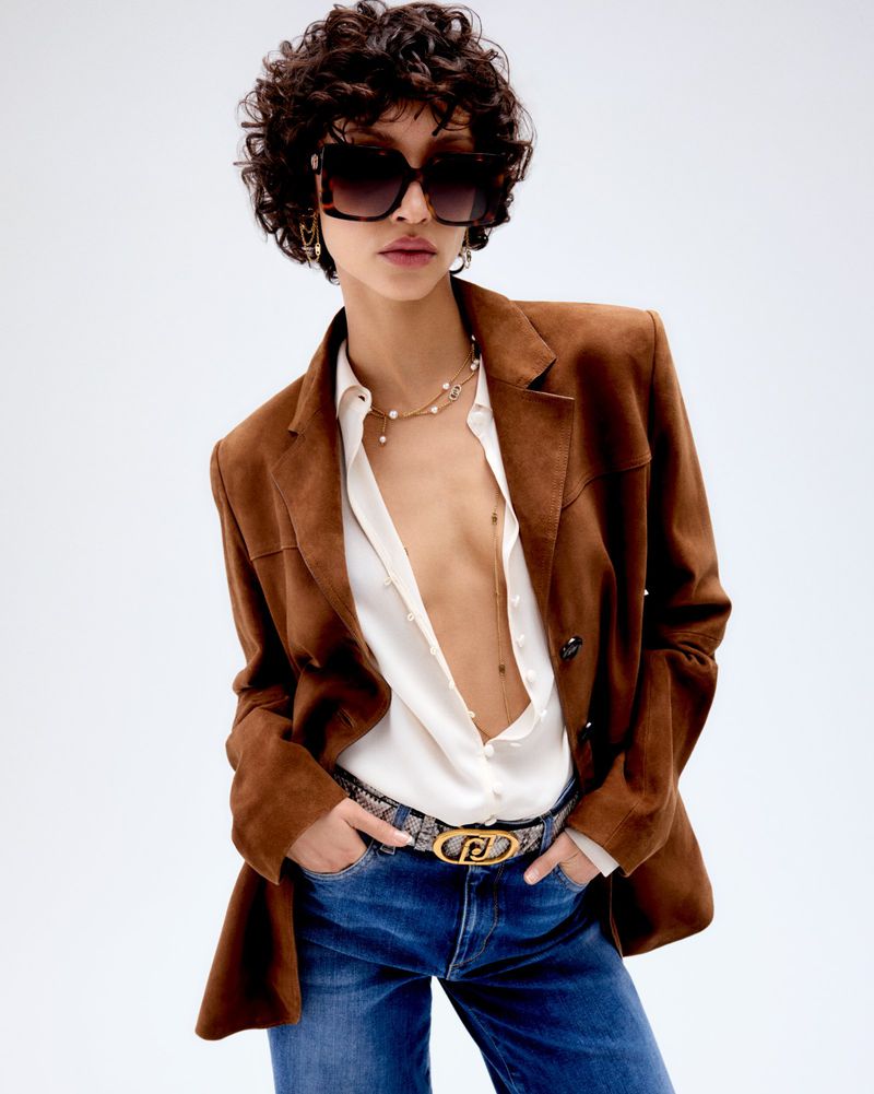 A young woman poses in a brown suede blazer, unbuttoned white top, blue jeans, and sunglasses against a plain studio backdrop.