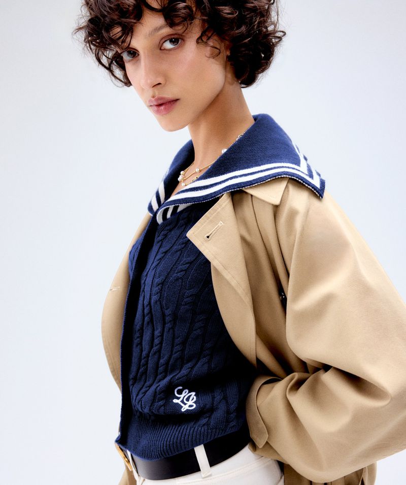 Fashionable adult female posing in a stylish navy cable-knit sweater with a sailor collar and tan trench coat against a white backdrop.