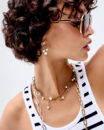 Model wearing gold aviator sunglasses, layered pearl necklaces, and a striped tank top against a white background.