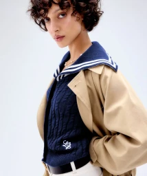 Model with short curly dark hair wearing a blue sailor collar sweater under a beige trench coat against a white backdrop.