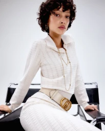 Model with short dark curly hair wearing a white textured polo shirt and skirt, seated on a black leather chair against a white background.