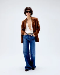 Model wearing a brown blazer, open white shirt, wide-leg blue jeans, and sunglasses against a plain white studio backdrop.