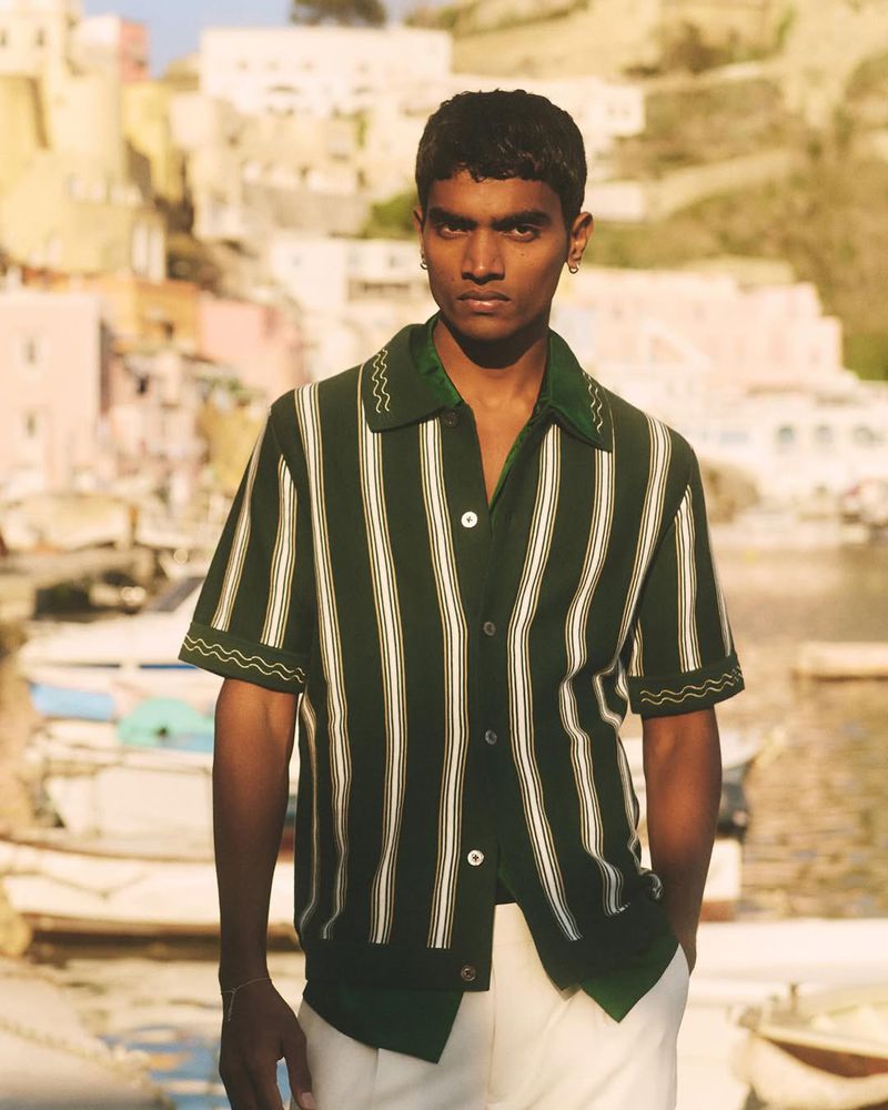 Model Deepak Gupta in a green and white striped knit shirt and white trousers standing before a coastal harbor.