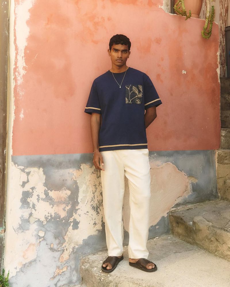 Model Deepak Gupta in navy t-shirt and white trousers standing against weathered pink wall, photographed by Olivier Yoan for CHÉ Studios High Summer 26