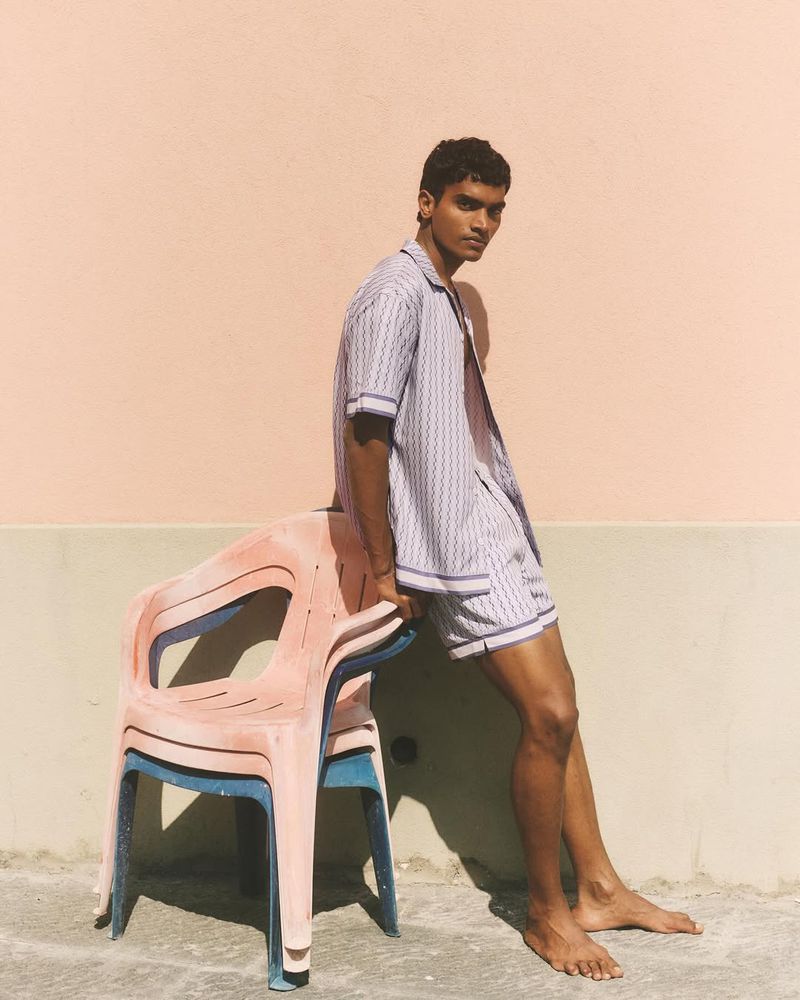 Model Deepak Gupta leaning on stacked plastic chairs wearing a matching patterned shirt and shorts against a warm wall.