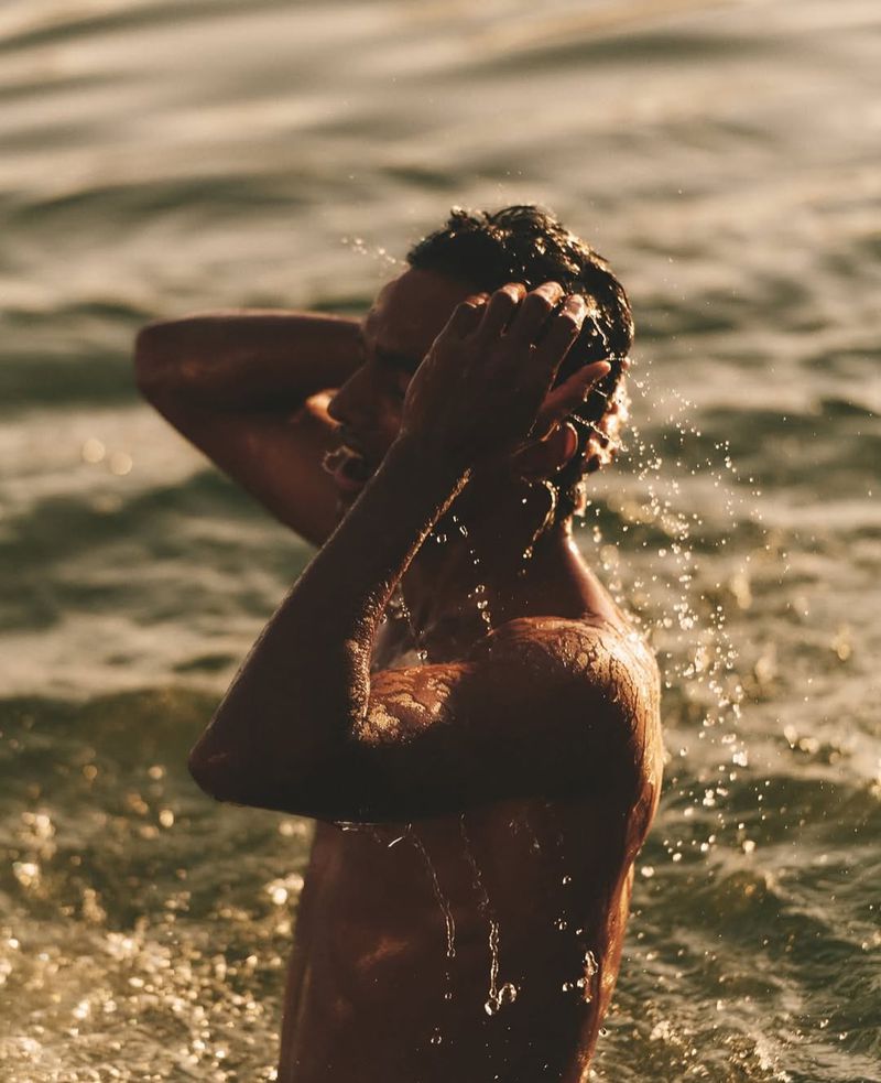 Model Deepak Gupta running his hands through wet hair while submerged in glistening ocean water during golden hour.