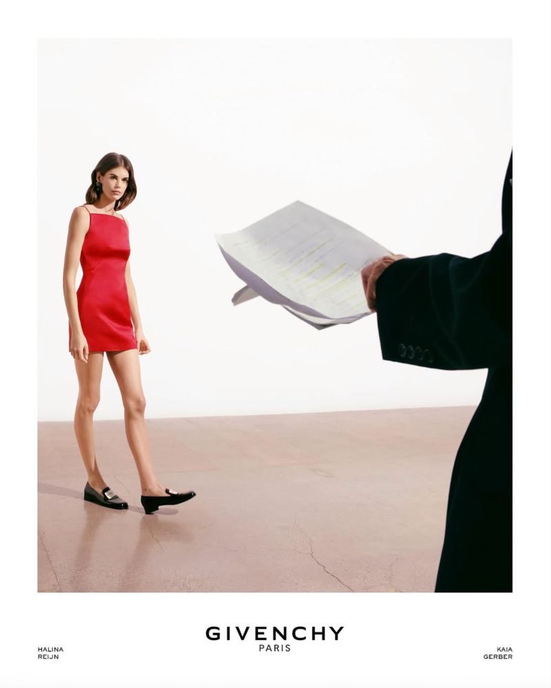 A fashion shot features a slender woman in a red dress and black loafers standing on a cream surface next to a suited person.