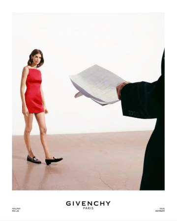 A fashion shot features a slender woman in a red dress and black loafers standing on a cream surface next to a suited person.