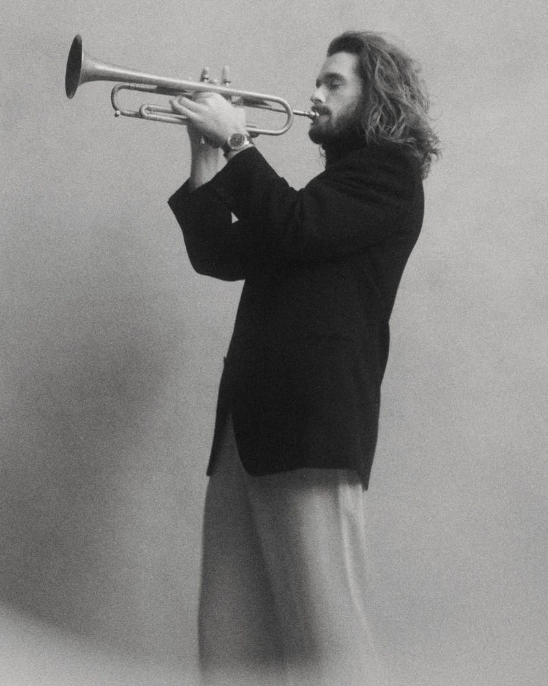 Man with long wavy hair wearing a black blazer and wristwatch playing a trumpet against a textured grey backdrop.