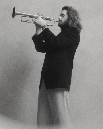 Man with long wavy hair wearing a black blazer and wristwatch playing a trumpet against a textured grey backdrop.