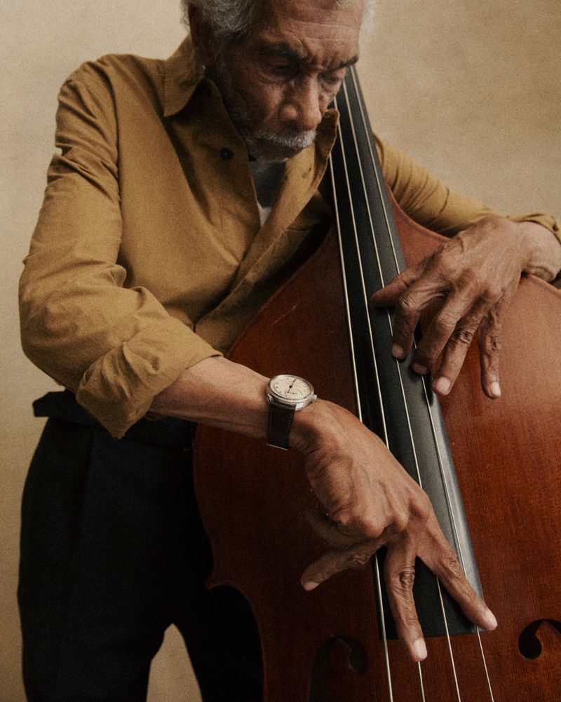 Elderly man playing a double bass wearing a mustard button-down shirt and a wristwatch for the Urban Jürgensen Spring 2026 Campaign.
