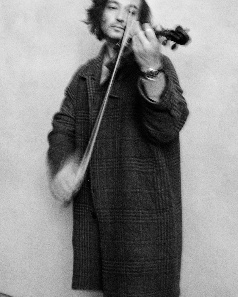 A man wearing a plaid overcoat plays the violin against a plain studio background in a grainy black and white portrait.