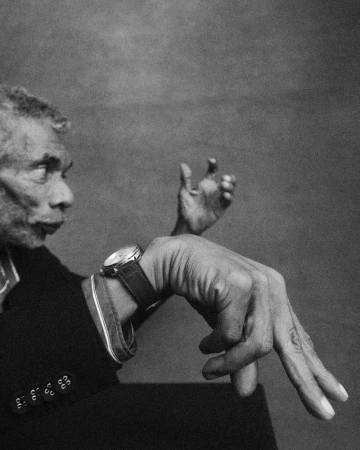 Black and white profile of an elderly man wearing a luxury wristwatch while gesturing with his hands against a studio backdrop.