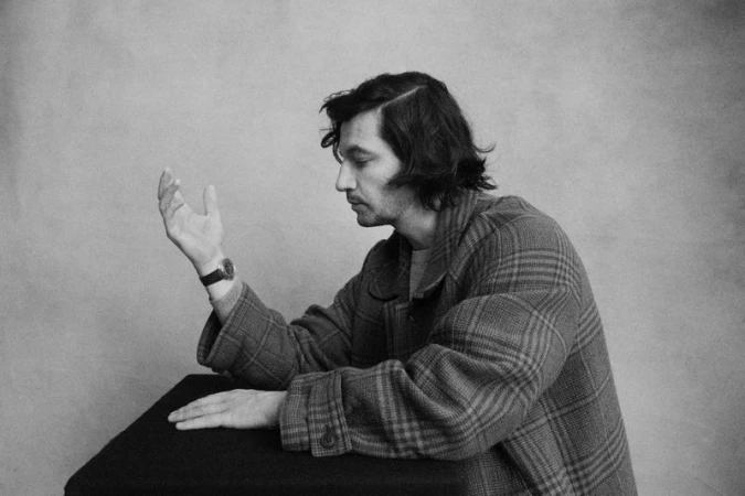 Black and white profile portrait of a man wearing a plaid coat and wristwatch, gesturing with an open hand at a dark table.