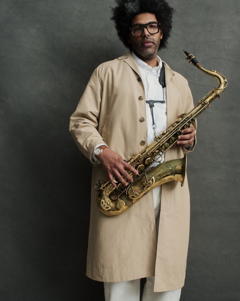 A man in a beige trench coat and glasses holding a vintage brass saxophone against a textured grey studio backdrop.