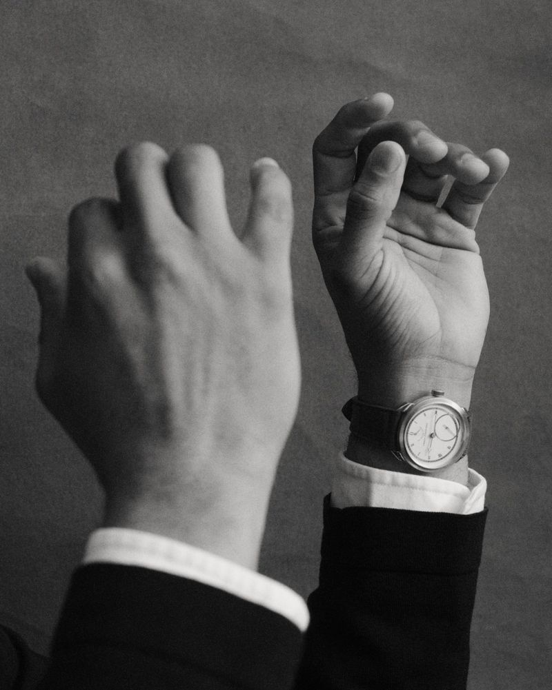 Monochrome close-up of a person's hands and wrist wearing a luxury dress watch with a dark suit and white cuff.