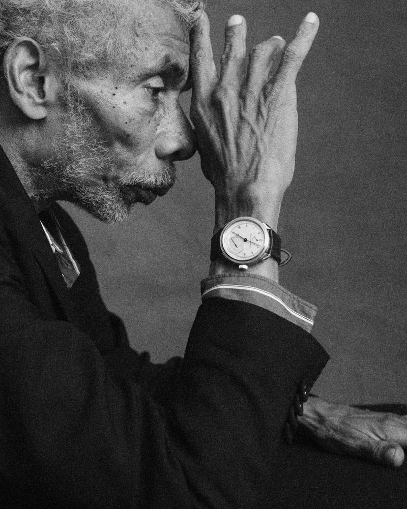 Black and white profile portrait of an elderly man resting his hand on his head while wearing a luxury wristwatch and dark blazer.