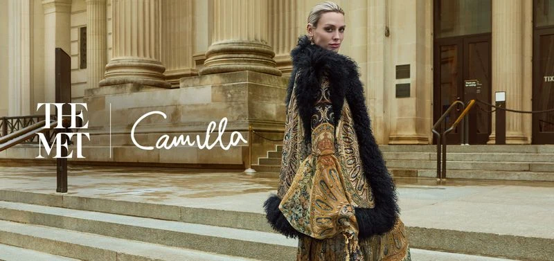 Woman wearing ornate patterned coat with black fur trim on classical stone building steps for The Met x CAMILLA campaign.