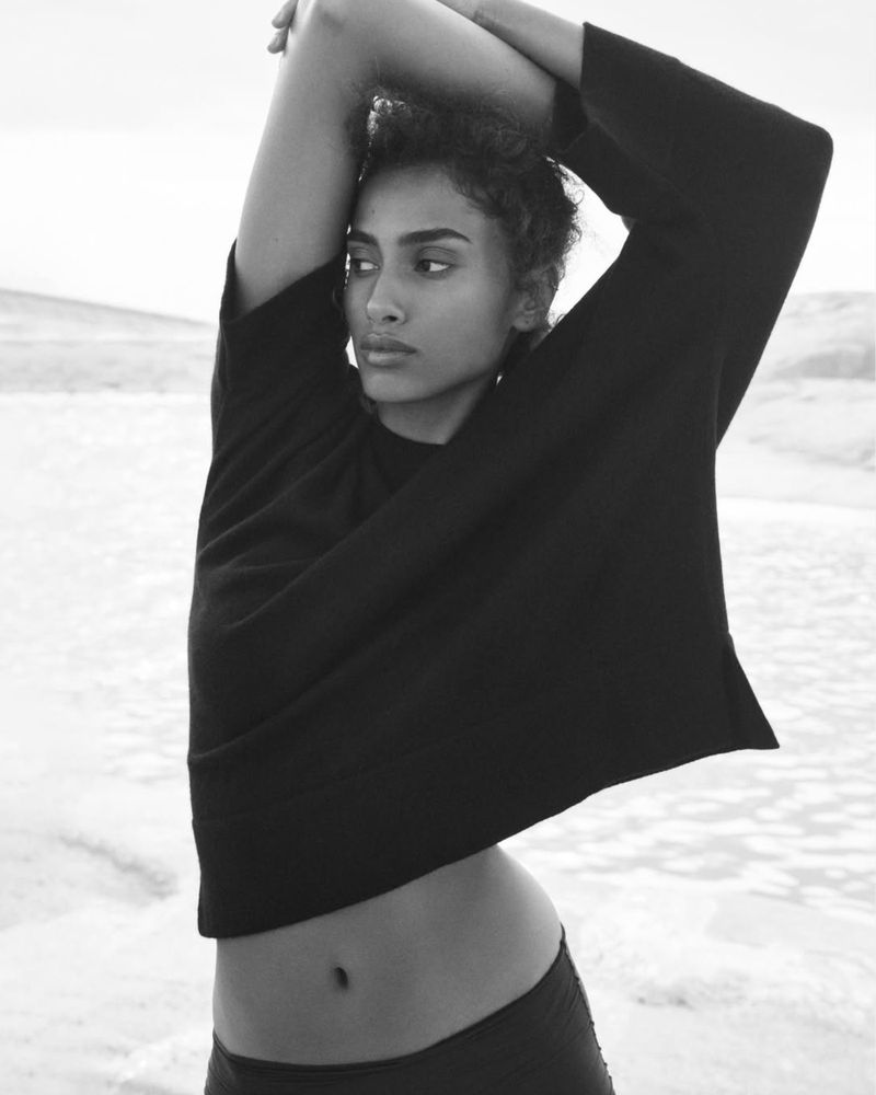 Portrait of a woman in a black top with her arms raised, set against a blurred background of sky and light-colored terrain.