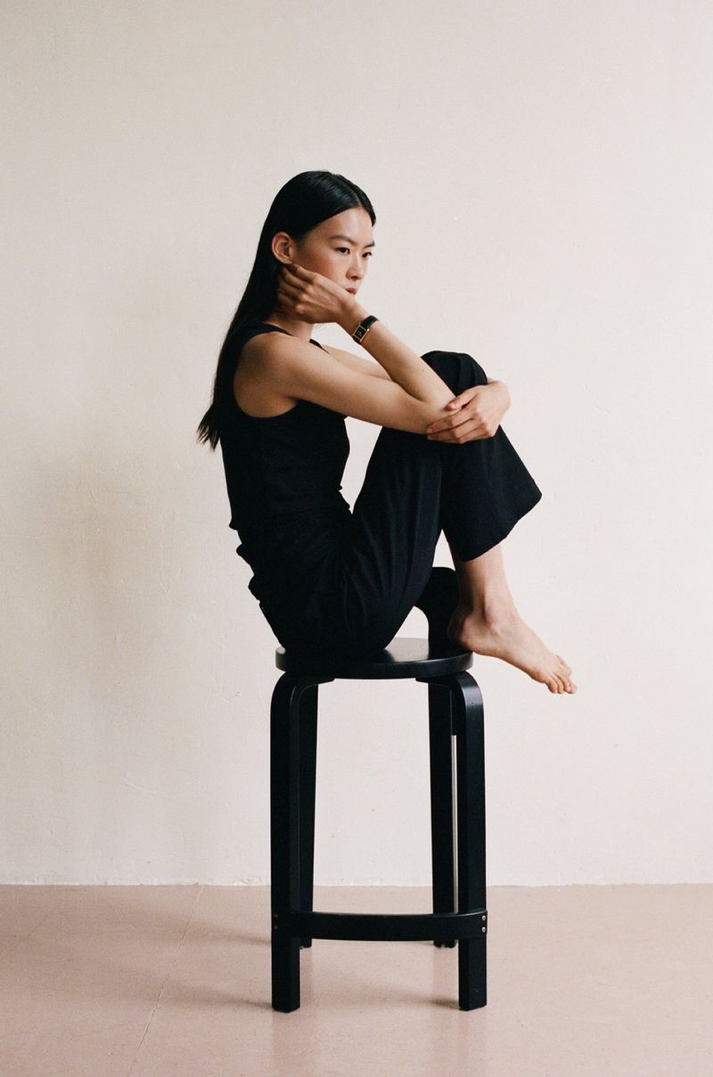 East Asian woman in black top and trousers seated barefoot on a black stool with hand to face in a studio setting.