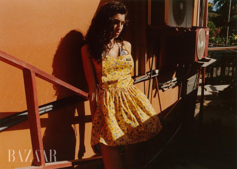 Model Mariana Goncalves in a printed yellow apron dress standing beside an orange exterior wall in direct sunlight.