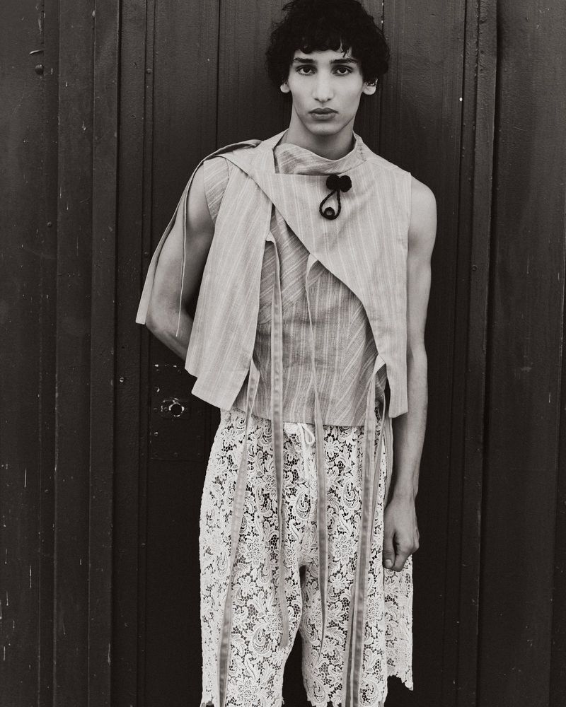 Monochrome portrait of a male model wearing an asymmetric sleeveless top and patterned shorts against a dark vertical-paneled background.