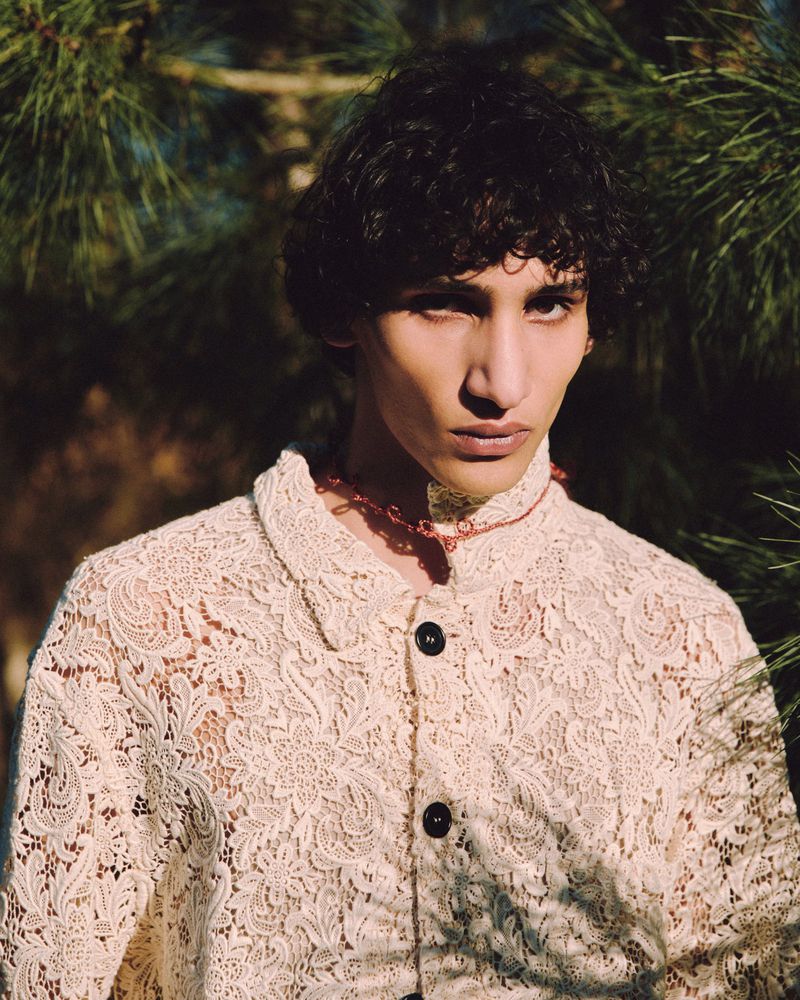 Portrait of a male model wearing a cream patterned lace button-up shirt outdoors against a pine tree background