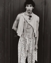 Monochrome portrait of a male model wearing an asymmetric sleeveless top and patterned shorts against a dark vertical-paneled background.