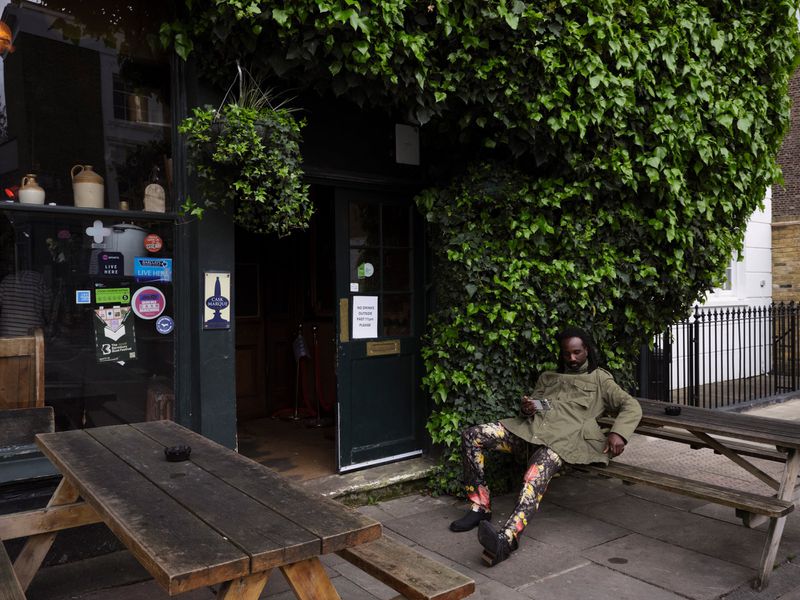 Man in an olive green jacket and patterned trousers seated on a wooden picnic bench outside an ivy-covered pub entrance.