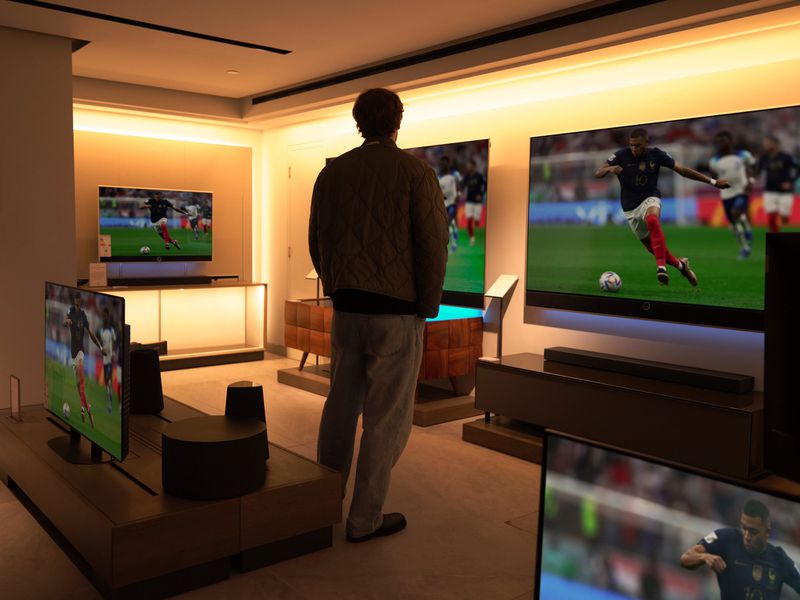 A man in a quilted jacket stands with his back to the camera, viewing multiple television screens playing a soccer match in a showroom.