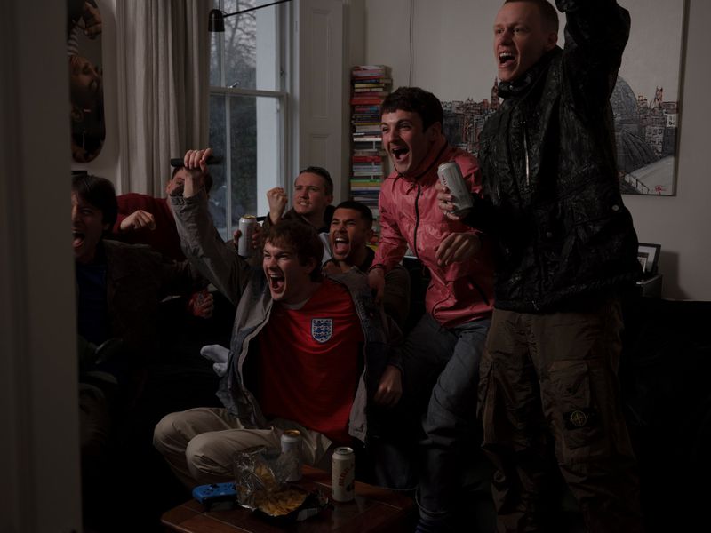 Group of men cheering in a living room, one wearing a red England football shirt and another in a black jacket.