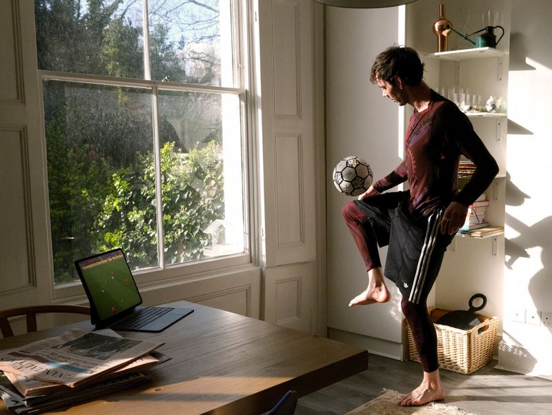 A barefoot man balancing a soccer ball on his knee in a sunlit room beside a dining table with a laptop showing a football match.