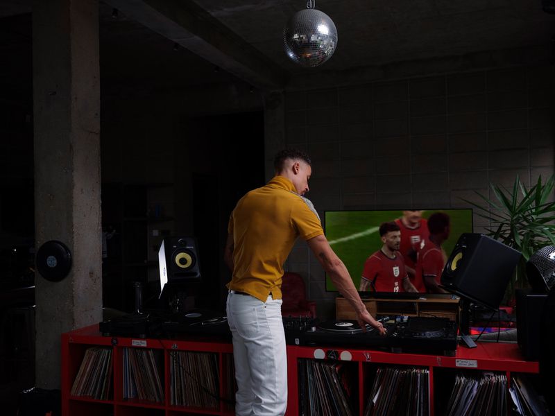 Man in a yellow polo shirt and white trousers DJing on turntables in a dim room with a television playing soccer in the background.