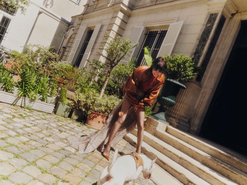 Woman in a leather jacket and long skirt stands on stone steps with a white dog in front of a building with green plants.