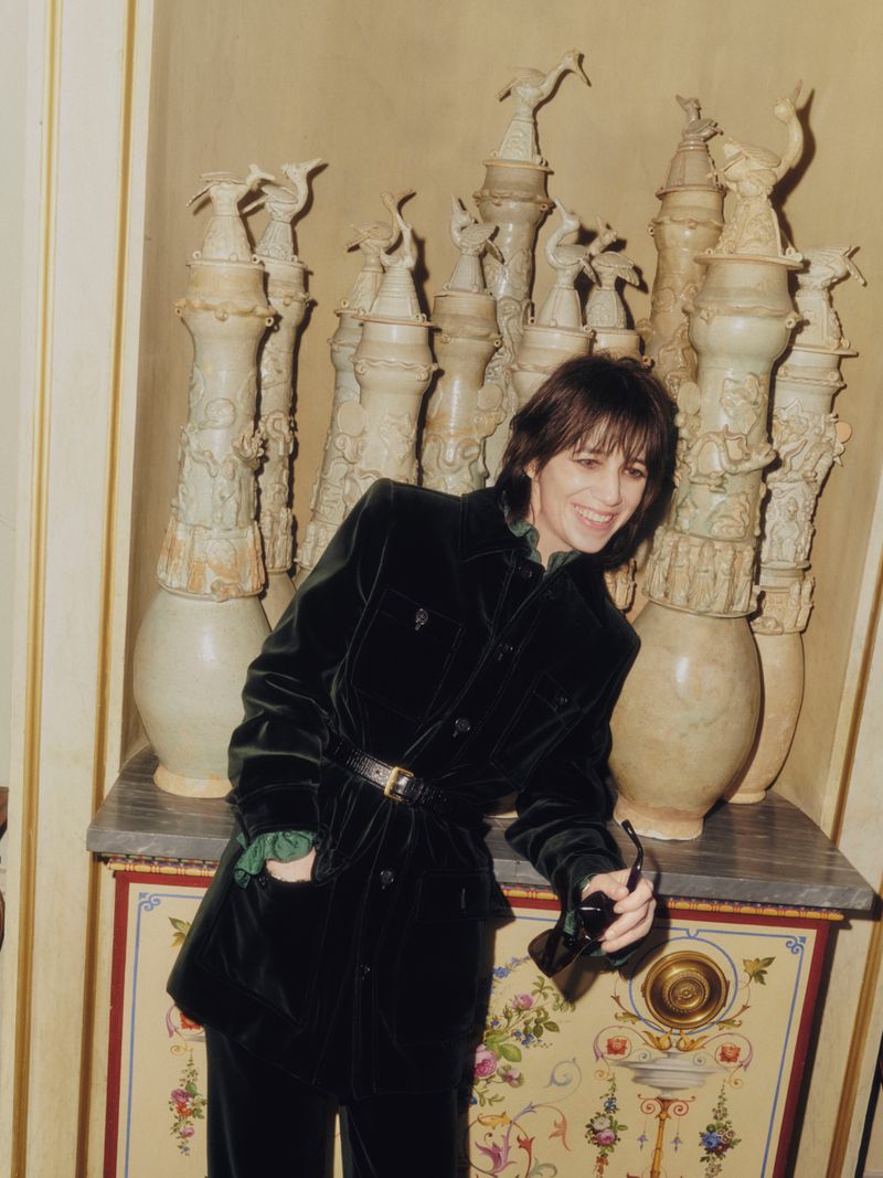 Smiling woman in green velvet suit stands indoors near decorative vases and furniture, holding sunglasses, lit by soft lighting.