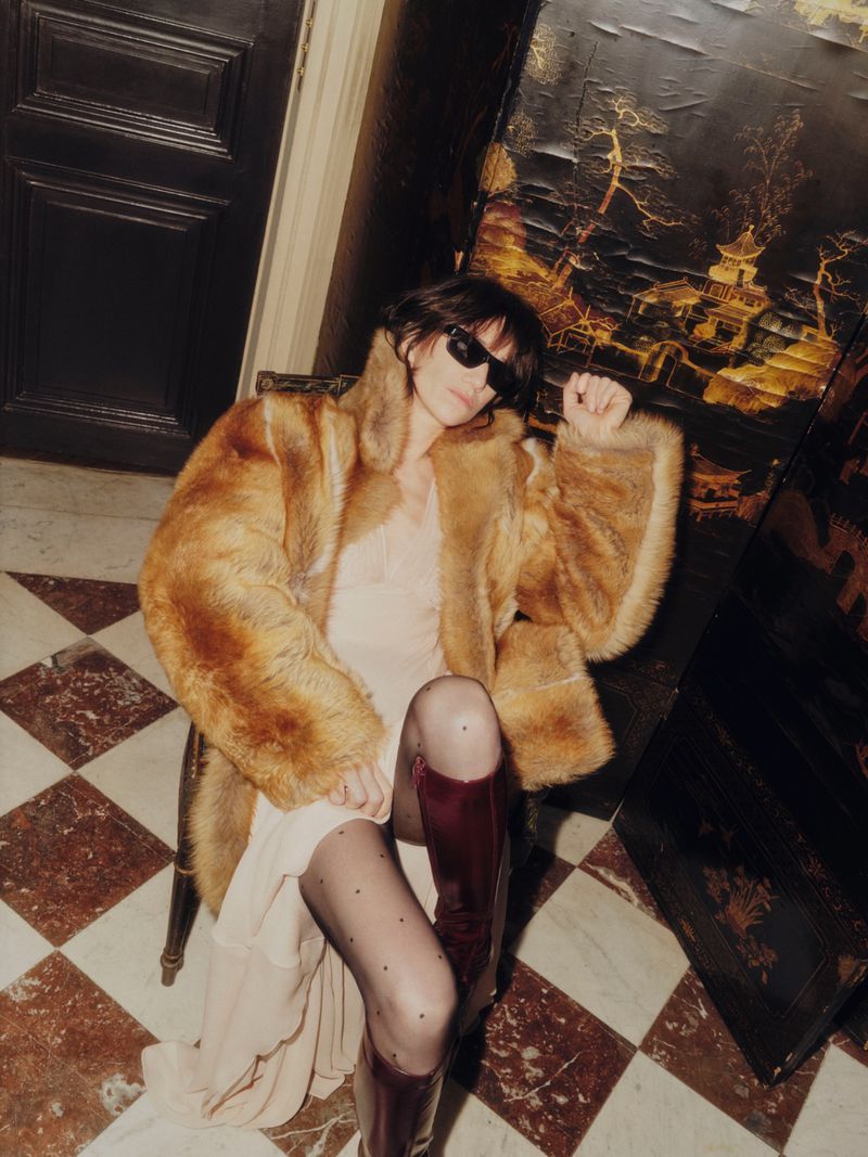 Woman sits in a chair wearing a fur coat, sunglasses, sheer dress, patterned stockings, and boots, beside a black decorative panel.