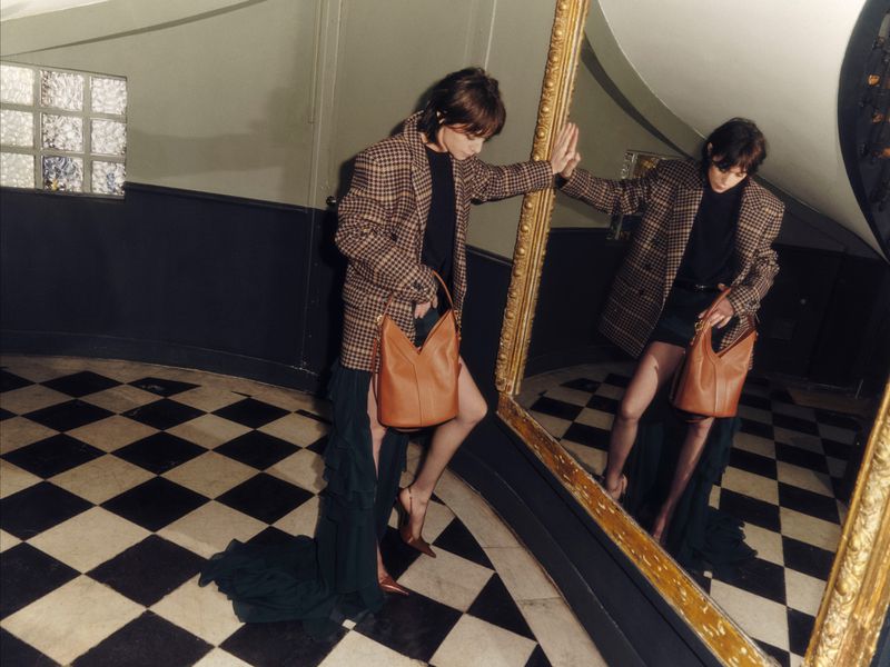 Woman poses near mirror in a plaid blazer with a brown leather handbag. Checkerboard floor, dark walls and natural lighting.