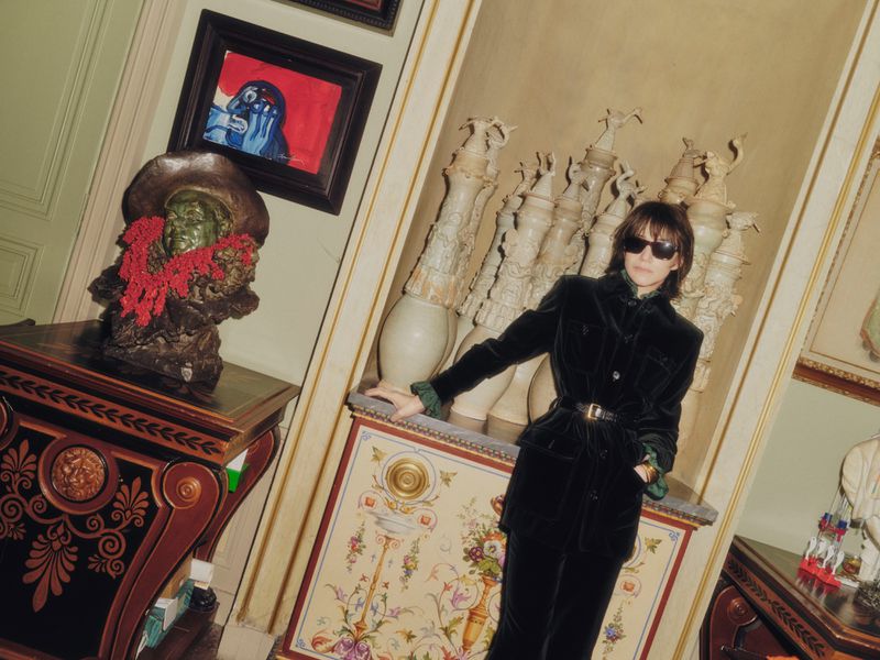 A woman in a velvet suit poses in an ornate room with sculpture, paintings, and antique furniture with cream and dark wood accents.
