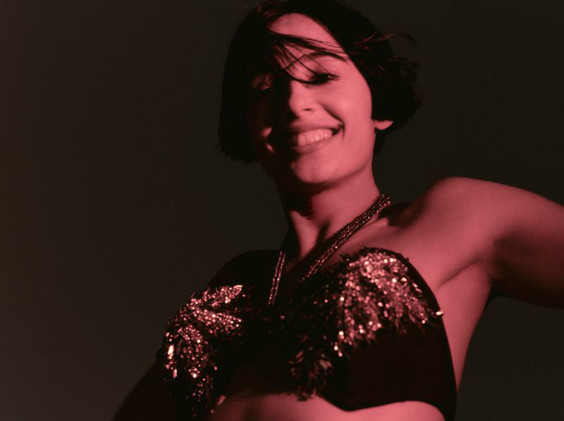 Naomi Scott smiling in a sequined embellished black strapless top under deep red lighting against a dark background.