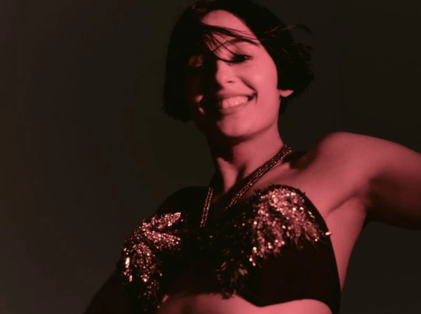 Naomi Scott smiling in a sequined embellished black strapless top under deep red lighting against a dark background.