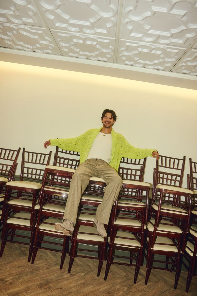 Matt Cornett wearing a lime green cardigan and tan pants sits atop a tall stack of banquet chairs indoors.