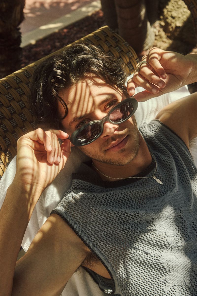 Young man reclining in a woven lounger wearing sunglasses and a textured grey knit top with natural shadow patterns