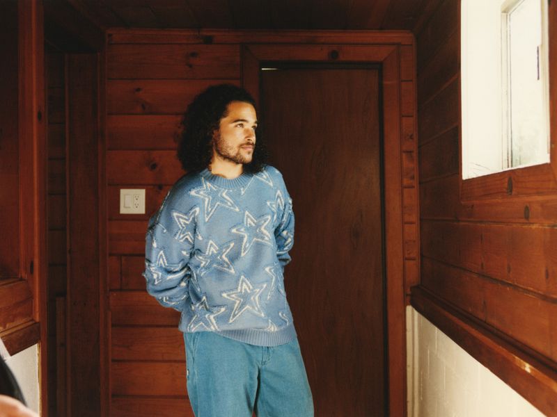 Actor Jalen Thomas Brooks wearing a blue star-patterned knit sweater and blue trousers in a wood-paneled room.