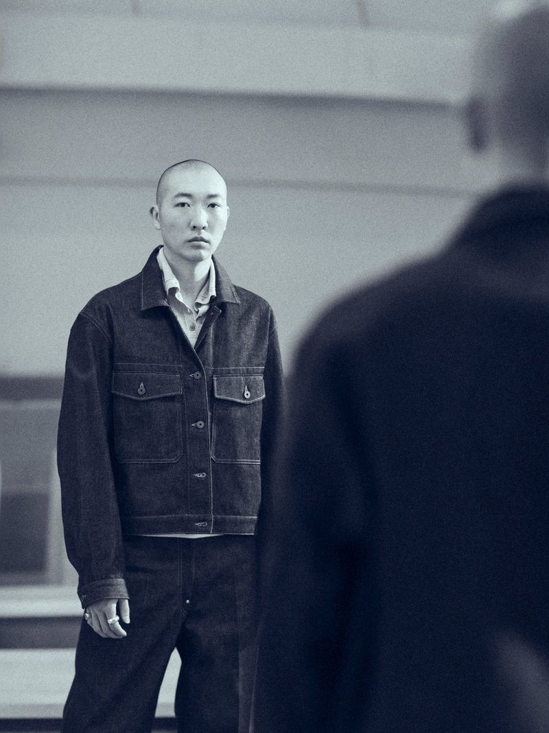 A bald person wearing a dark denim jacket and matching trousers looking towards the camera in an industrial interior setting.